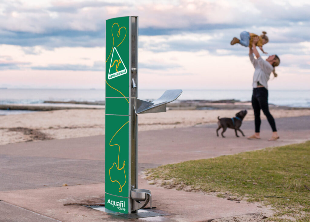 Aquafil by Civiq, Australian Made Drinking Fountain shown placed ona beach south of Sydney A mum lifts her child in the airm and the dog plays.