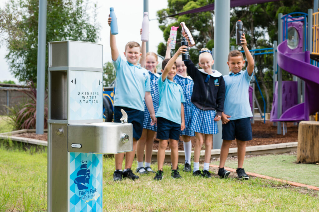 Students raising their drinking water bottles with an Aquafil Drinking Fountain in the background