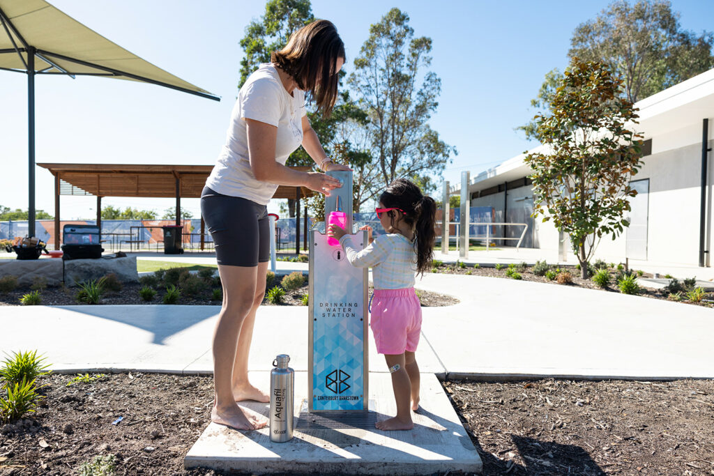 A mother and her daughter drinking on an Aquafil Bold Drinking Fountain for Open Spaces and Playgrounds