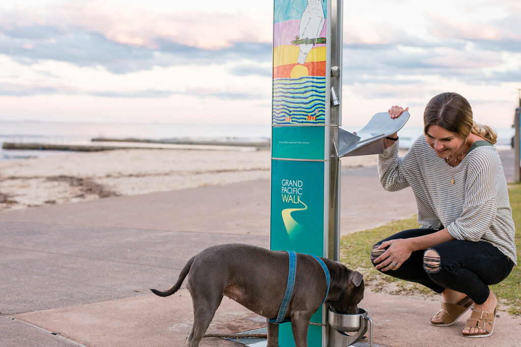 Woman watching her dog drinking on an Aquafil Drinking Water Station installed near a beach shore in Wollongong City