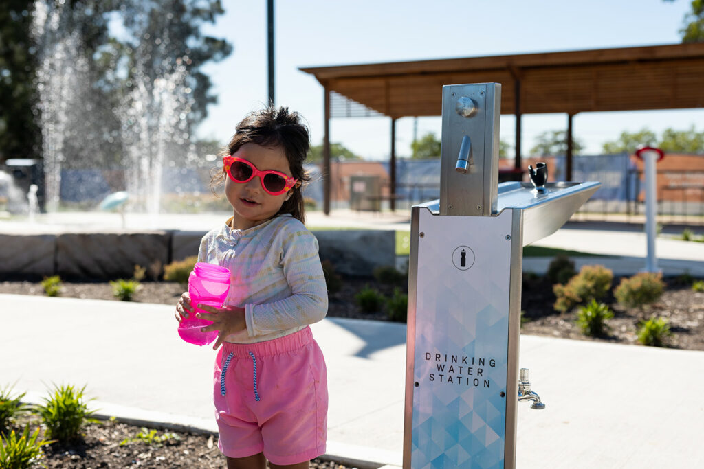 A delighted child holds her water bottle beside the Aquafil bold drinking fountain and water bottle refilling station, ready to quench her thirst