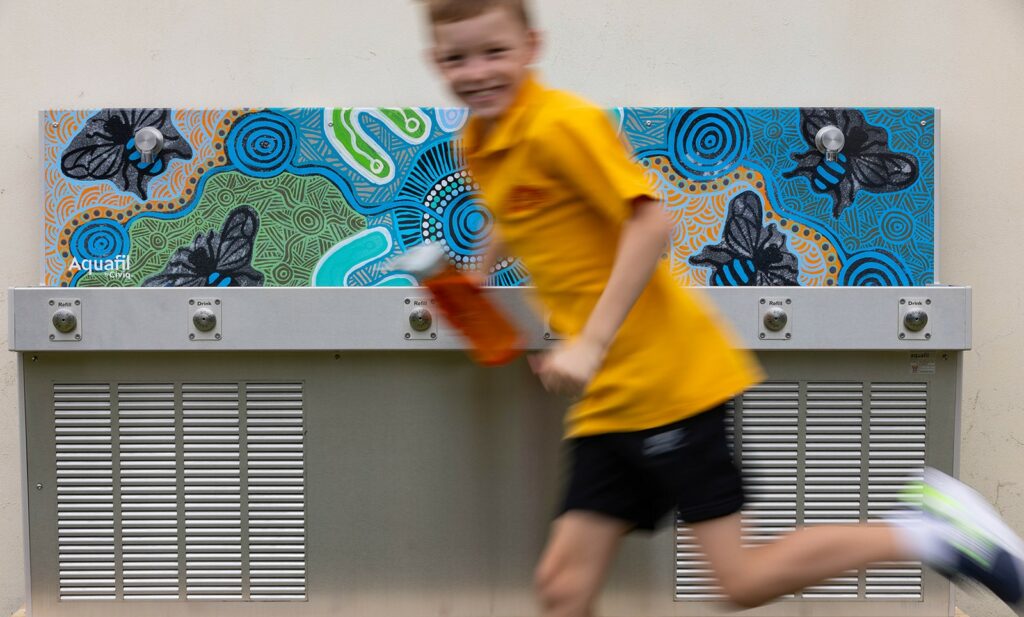A joyful student runs past an Aquafil Hydrobank by Civiq, clutching his water bottle with a grin. The school drinking trough, adorned with Luke Penrith's vibrant Aboriginal artwork, adds a burst of color to the scene, blending art, hydration, and energy in motion.