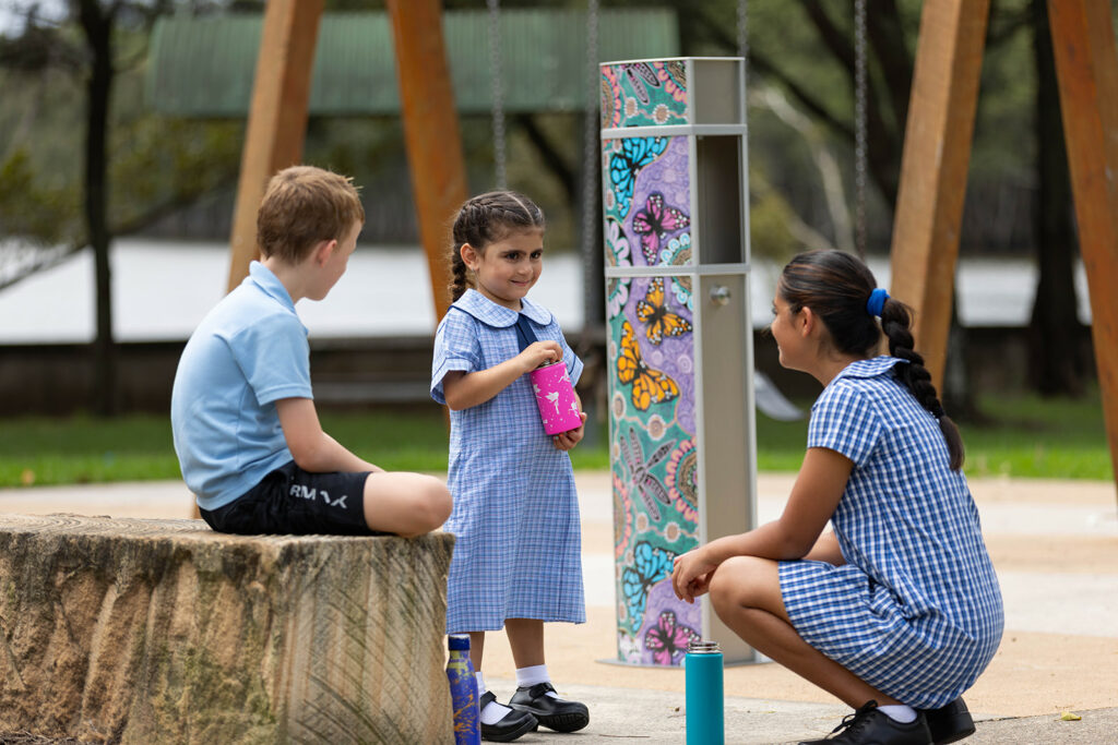 Students stand proudly with their eco-friendly water bottles, poised beside the vibrant Aquafil School water bottle refilling station adorned with stunning butterfly Aboriginal artwork.