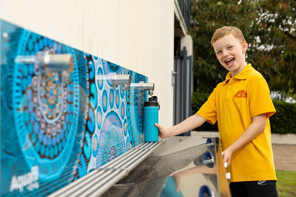 A student refills his water bottle at the Civiq Aquafil Hydrobank School Drinking Trough, embellished with vibrant Aboriginal artwork by Luke Penrith. Celebrating culture and hydration in one vibrant scene.